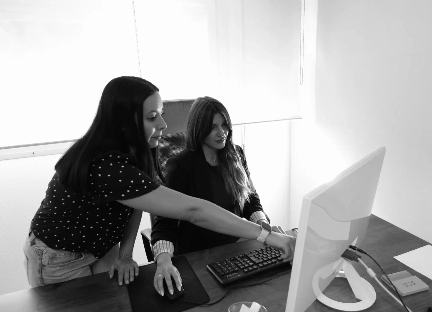 Two colleagues working together at a desktop computer in an office, in black and white
