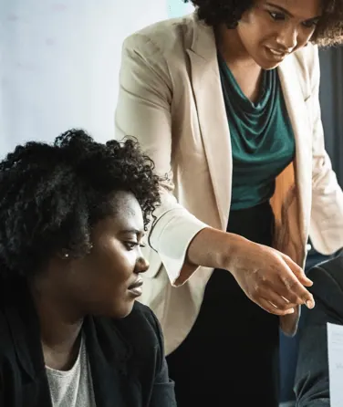Two women in an office: one stands in a beige blazer and points, the other sits and follows with her eyes